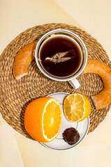 A cup of tea with a tea bag, a saucer with citrus fruits and candy, next to a bagel with poppy...