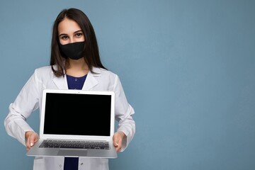 Side profile shot of Young brunet woman wearing medical white coat and black mask holding laptop and looking at camera isolated on blue wall background