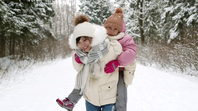 Locked-down shot of young Asian woman carrying happy daughter on back in white snowy winter road in forest