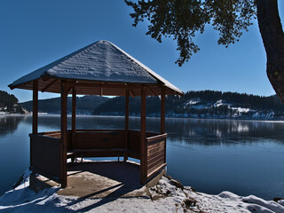 Snow-covered wooden pavillon on the shore of frozen Schluchsee lake, a popular tourist destination in Black Forest mountain range, Germany, in winter season on sunny day with blue sky. - Powered by Adobe