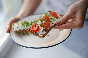 Woman hands holding plate with rye crisp bread with creamy vegetarian cheese tofu, cherry tomato...