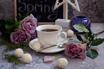 Women's holidays concept. White cup with tea, coconut cakes, lilac roses and the inscription Spring on a gray background.