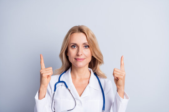 Photo of mature doctor woman with blonde hair and white coat point fingers copyspace isolated on grey color background