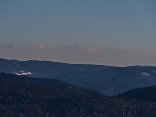 Stunning aerial view of the southern Black Forest hills in winter season with snow-covered landscape viewed from Schauinsland peak, Germany with the silhouettes of the majestic Alps in background.