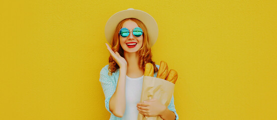 Portrait of happy smiling young woman holding grocery shopping paper bag with long white bread baguette on a yellow background