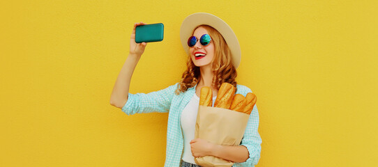 Portrait of happy smiling woman taking a selfie picture by phone holding grocery shopping paper bag with long white bread baguette on a yellow background - Powered by Adobe