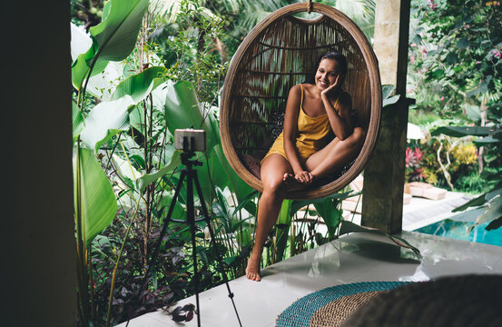 Smiling female chilling on wicker hanging chair and taking selfie on smartphone
