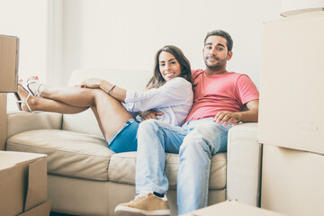 Happy Latin young couple sitting on couch among cardboard boxes in new house, looking at camera and smiling. Moving or relocation concept