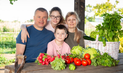 Portrait of happy family with two kids posing in garden with fresh harvest of vegetables and greens