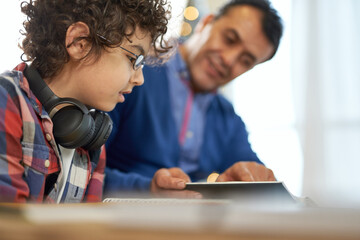 Enthusiastic latin boy wearing glasses looking focused while sitting at the desk together with his father and doing homework during remote learning at home - Powered by Adobe