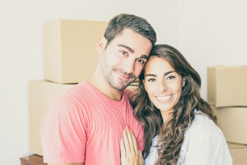 Happy beautiful Hispanic couple standing among carton boxes in their new flat, hugging and looking at camera. Medium shot. New home and real estate concept