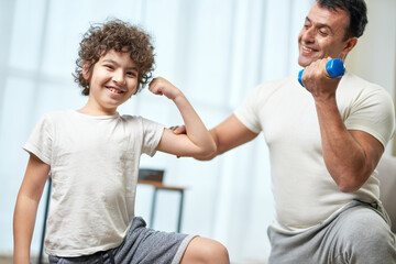 Fototapeta premium Fit boys. Little hispanic boy child smiling at camera showing his biceps while having workout together with his father, exercising at home