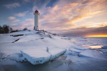 Graceful white lighthouse staying on the rocky coast covered with snow and ice at sunset. Russia,...