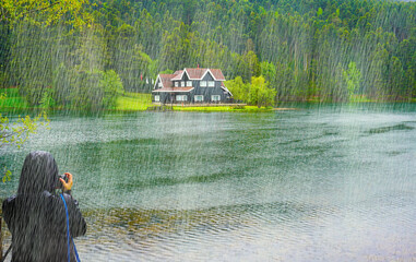 Woman photographer taking photo of impressive lake house. It is raining. House on the shore of Cennet Gol (Paradise Lake), Bolu. Reflected on water. Country side, forest, farm field, lake with house.  © Akin Ozcan