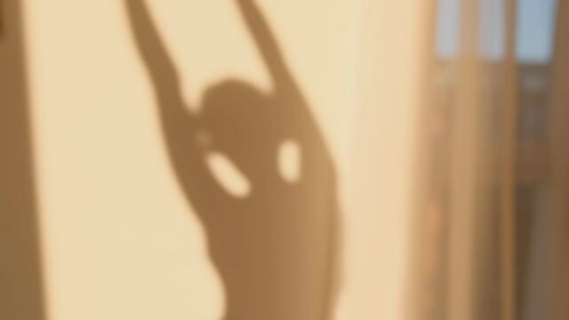 Shadows silhouette woman waking up doing stretching and morning exercises at home sunset or dawn in the rays the sun reflected on wall. Golden hour through window curtains. Blurry and out of focus.