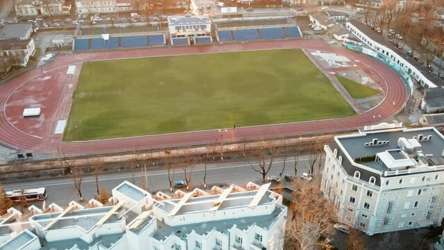 Aerial drone view of a stadium in Chisinau. Green field, running tracks with a man, tribunes, buildings nearby, roads with moving cars and bare trees. Moldova