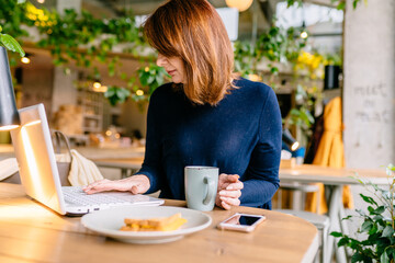 Red haired 50 age plus woman in blue sweater sitting in restaurant or coffee shop and working with laptop.
