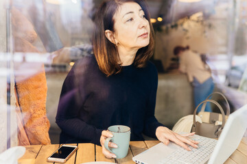 View through the window red haired 50 age plus dreamy woman in blue sweater sitting in restaurant or coffee shop and working with laptop. Female sits behind the glass of a cafe.