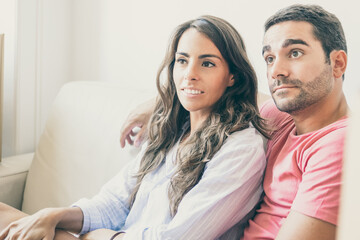 Positive focused Latin couple sitting on couch among carton boxes in new apartment, looking away. Moving or new home concept