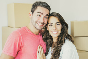 Happy young couple enjoying moving into new flat, standing among carton boxes, hugging and looking at camera. Medium shot. New home and real estate concept