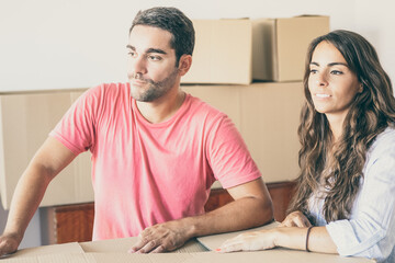 Pensive young man and woman standing among carton boxes in apartment, looking away. Medium shot....