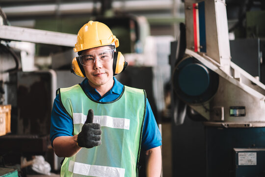 Work At Factory.Asian Worker Man  Standing With Showing Thumbs Up To Look At The Camera In Safety Work Wear With Yellow Helmet.in Factory Workshop Industry Machine Professional