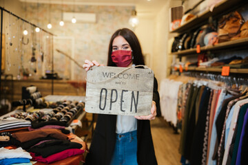 Portrait of a beautiful young clothing store sales assistant holding the "Welcome we're open" sign at the business entrance wearing a face mask during the Coronavirus Covid-19 pandemic