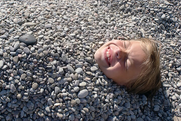 A little girl lies on the beach under the pebbles. A playful baby is buried in the pebbles on the beach, only her head is visible. A child on the beach. The concept of a happy childhood, a fun holiday