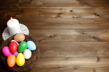 Burning white wax candle on openwork napkin, birds nest with multicolored easter eggs on dark wooden planks desk with copy space. Easter holiday banner, card, poster, voucher, invitation template