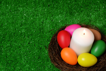 Burning white wax candle in birds nest surrounded by multi colored easter eggs on green grass lawn and wooden panks background top closeup view selective focus with copy space