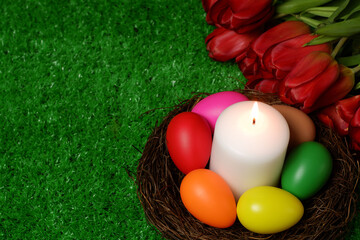 Burning white wax candle in birds nest surrounded by multi colored easter eggs, bouquet of red tulip flowers on green grass lawn background top closeup view selective focus