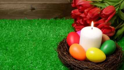 Burning white wax candle in birds nest surrounded by multi colored easter eggs, bouquet of red tulip flowers on green grass lawn and wooden panks background side closeup view selective focus