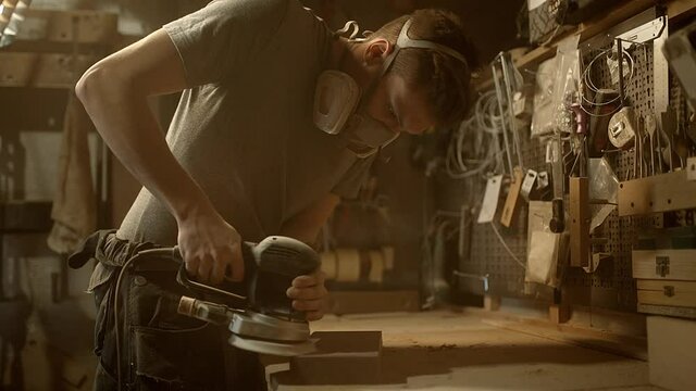 A young carpenter grinds a piece of wood in a half mask respirator in his workshop. High quality 4k footage.