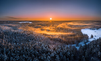 Warm sunrise over snowy countryside landscape. Pine forest covered in fog waves. Drone aerial view.