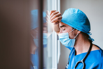 Doctor in mask and uniform leaning against a window looking exhausted
