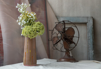 Bouquet of green and white flowers in Handmade ceramic vase and Vintage fan on white textured table cloth with old cement wall. Home decor, Selective focus.