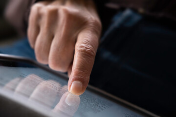 Close-up of finger and hand on the touchscreen of an iPad tablet with dark background and reflection of the fingers in the screen. Focus on the index finger