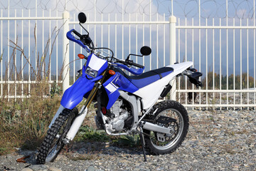 Blue and white enduro motorcycle on the background of a fence with barbed wire. An off-road motorcycle is parked on a rocky beach.