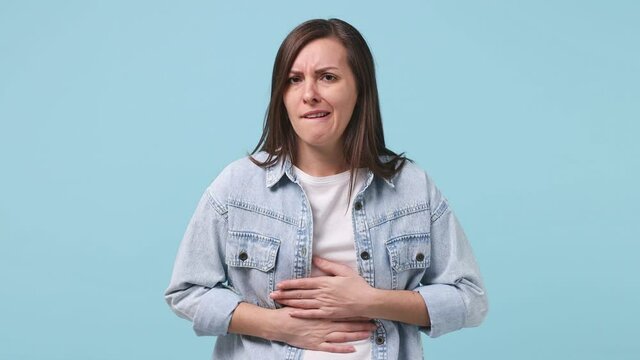 Sick ill sad young woman 30s years old in denim jacket white t-shirt put hands on abdomen suffering from stomach-ache griping bellyache period feel bad seedy isolated on pastel blue background studio