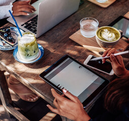 Warm portrait of a asian woman she sits at table with coffee and water cups and works on her tablet outside in sunny day.