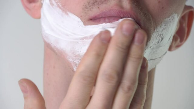 A young man applies foam or shaving gel close-up in the bathroom. Facial skin care, shaving facial hair, stubble, mustache and beard. Unshaven guy