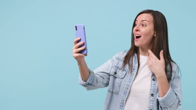 Woman in denim jacket white t-shirt get video call using mobile cell phone doing selfie videoconference talk conducting pleasant conversation greet with hand isolated on pastel blue background studio