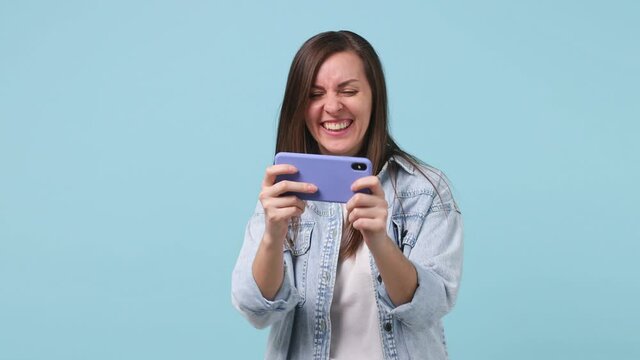 Gambling fun long hair young woman 20s years old in denim jacket white t-shirt using play racing on mobile cell phone hold gadget smartphone for pc video game isolated on pastel blue background studio