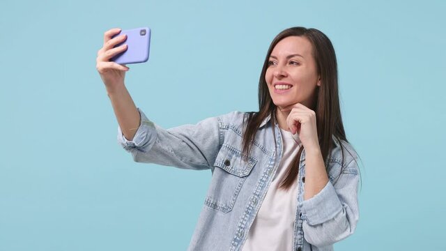 Smiling beautiful long hair brunette young woman 30s years old in denim jacket white t-shirt posing doing selfie shot on mobile phone isolated on pastel blue background studio People lifestyle concept