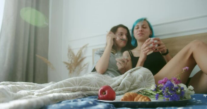Lesbian couple having romantic breakfast in bedroom. Young woman sharing food and kissing girlfriend with dyed hair while resting in comfortable bed in morning at home