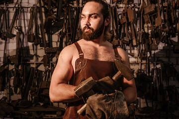 Muscular brutal blacksmith in a leather apron in the forge holding two hammers crossed in front of...