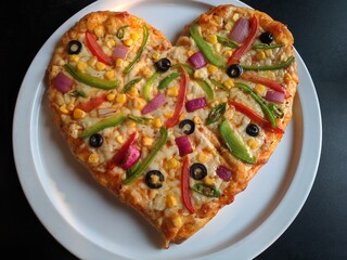 A delicious looking heart shaped vegetable cheese pizza served on a white round plate over a black surface table at a southeast Asian restaurant