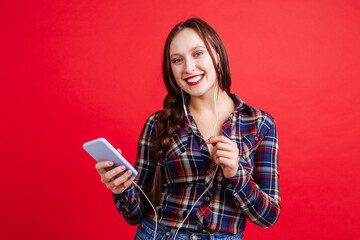 Beautiful girl dressed in casual style listening to music through headphones in the phone, on a red background