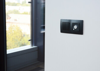 A close-up of a modern elegant black light switch and a climate, temperature control panel, the thermostat of a heater installed on a white wall in a room near a window.