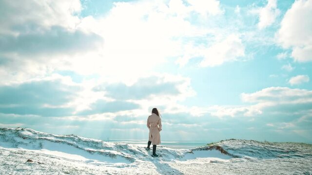 A young girl watching the ocean at the edge on top of a rocky seashore in 4K. A woman smartly dressed enjoying a lighthouse view during winter on a windy day, the wind blowing hair in slow motion.